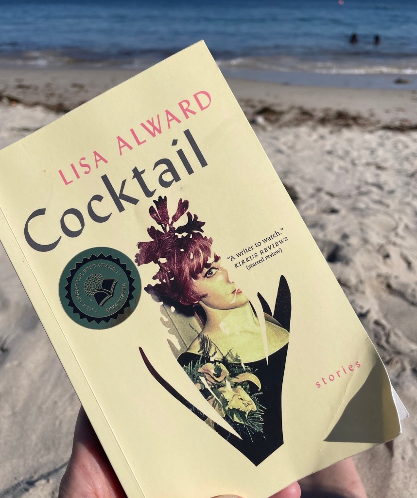 Image of front cover of "Cocktail" by Lisa Alward. The book is held at an angle by a hand and there is a sandy beach with gentle waves in the background. 